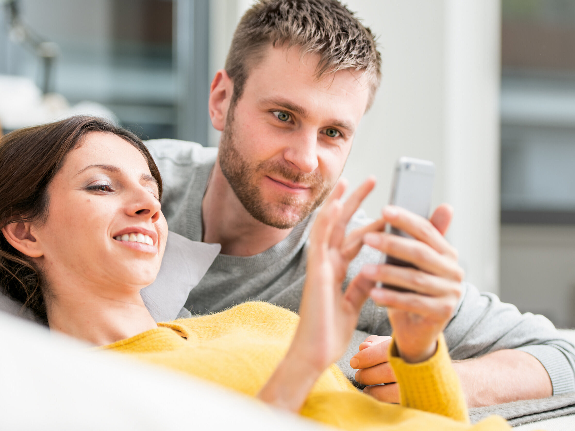 Raumluftregelierung über das Smartphone Frau liegend auf sofa betrachtet telefon, mann daneben betrachtet ebenfalls den telefondisplay